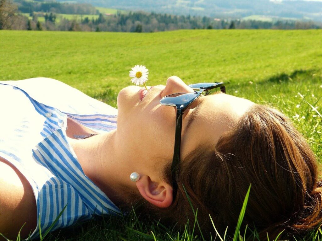 Chica en un prado sujetando una margarita con la boca y luciendo unas gafas de sol que la protegen de la radiación solar y ultravioleta tras su intervención de miopía.