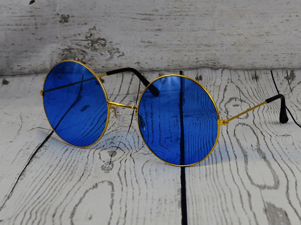 Una pareja de gafas de sol redondas con lentes azules. Las gafas tienen un marco de metal dorado y están colocadas sobre un fondo blanco.